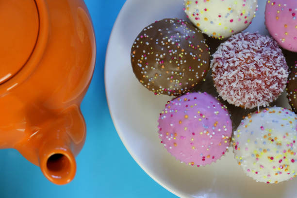 istock Image of multicolour homemade cupcakes / mini muffins cakes, vanilla, strawberry / raspberry, coconut, chocolate flavours with icing and rainbow sprinkles on round white plate with orange teapot, turquoise background for afternoon tea / children's birthda 1172159666