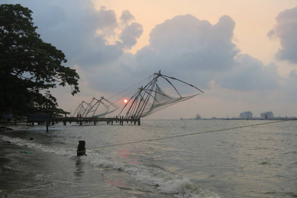 Stock photo of Chinese fishing nets at sunset in Fort Kochi beach in Cochin, Kerala, South India with local Indian fisherman catching tuna fish mackerel from port of Kochi harbour water in Indian Ocean / Arabian Sea, Cheena vala Chinese fishing nets Kochi photo imagen de las redes de pesca chinas al atardecer en la playa de fort kochi en cochin, kerala, al sur de la india con pescador indio local capturando caballa de atún desde el puerto de aguas del puerto de kochi en el océano indico / mar arábigo, cheena v