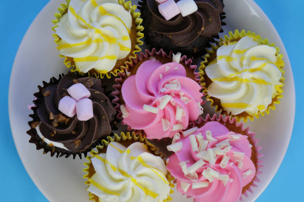 istock Image of small homemade cupcakes / mini muffins, rainbow multicolour icing piped cakes as flower swirl, marshmallow, pink strawberry, yellow lemon, cocoa chocolate icing cakes on round white plate for afternoon tea / children's birthday party food buffet 1172159800