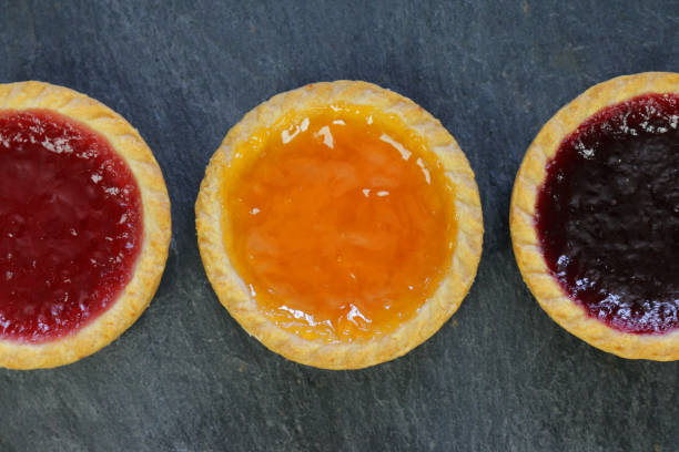 Image of small homemade jam tart pies with shortcrust pastry filled with strawberry, raspberry, apricot jam on top in a row with black  wooden background for afternoon tea / children's birthday party food buffet platter Stock photo of small homemade jam tart pies with shortcrust pastry filled with strawberry, raspberry, apricot jam on top in a row with black  wooden background for afternoon tea / children's birthday party food buffet platter bakewell photos stock pictures, royalty-free photos & images