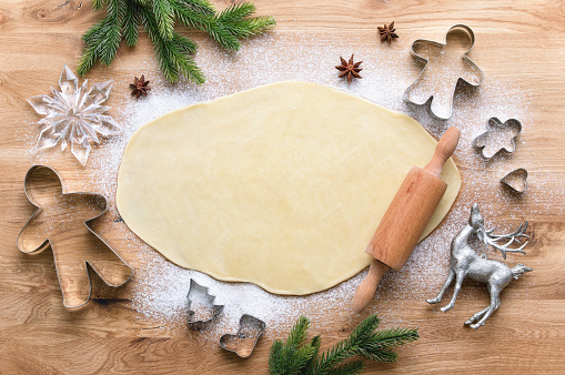 An overhead view of dough is rolled out on a wooden block cutting board with flour spread out and a wooden rolling pin. Ready to make Christmas Theme cookies with various metal cookie cutters for the holiday season.