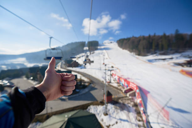 Human hand with thumb-up gesture on background of mountain ski-lift rope way on sunny winter day. Human hand with thumb-up gesture on background of mountain ski-lift rope way on sunny winter day. Moving cable cars, bright blue sky and dark spruce trees, view from above. Active lifestyle concept. thumbs up for skiing stock pictures, royalty-free photos & images