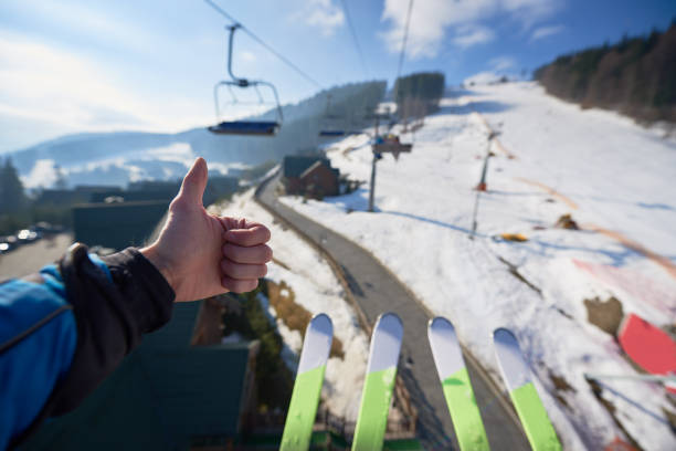 Hand with thumb-up gesture and skis on background of mountain ski-lift rope way on winter day. Winter landscape of ski resort. Human hand with thumb-up gesture and two pairs of skis on background of mountain ski-lift rope way on sunny winter day, top view. Sports and recreation concept. thumbs up for skiing stock pictures, royalty-free photos & images