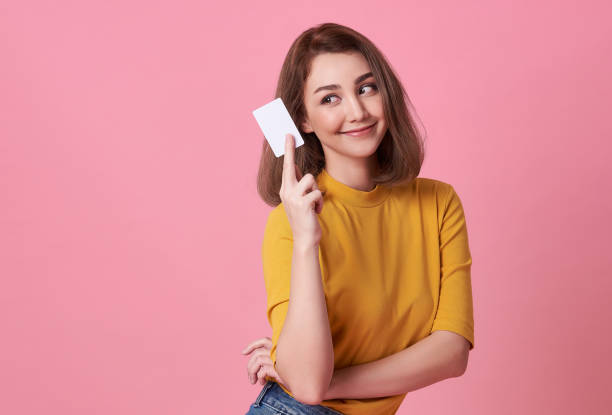 ritratto di una giovane donna in camicia gialla che mostra la carta di credito e guarda lontano lo spazio di copia isolato su sfondo rosa. - carta di credito foto e immagini stock