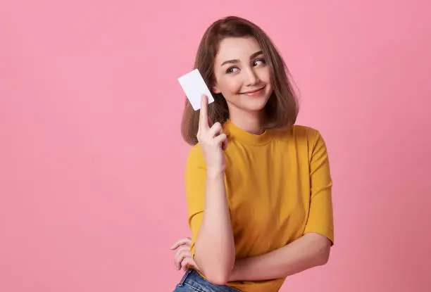 Portrait of a young woman in yellow shirt showing credit card and looking away at copy space isolated over pink background.