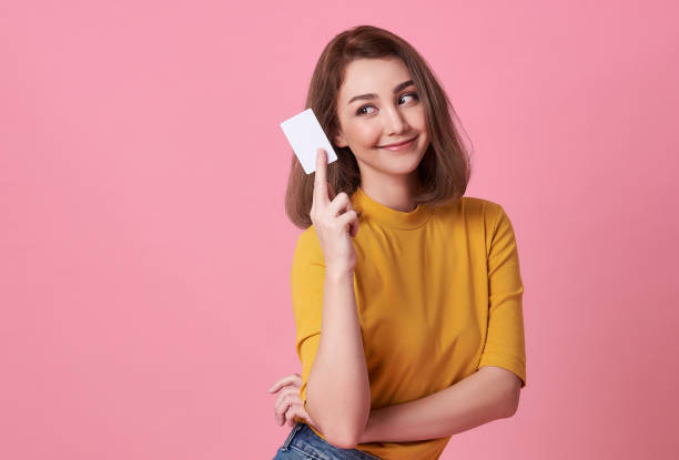 retrato de uma mulher nova na camisa amarela que mostra o cartão de crédito e que olha afastado no espaço da cópia isolado sobre o fundo cor-de-rosa. - holding money - fotografias e filmes do acervo