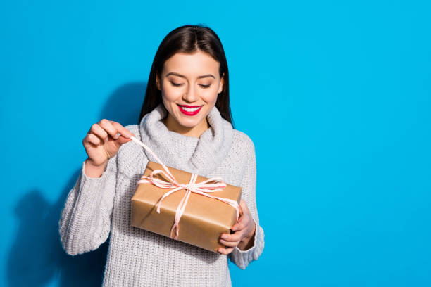 Portrait of cheerful lady pulling ribbon looking at package smiling wearing gray sweater isolated over blue background Portrait of cheerful lady pulling ribbon looking at package smiling wearing gray sweater isolated over blue background unwrapping stock pictures, royalty-free photos & images