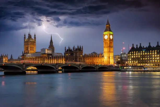 The Westminster Palace and Big Ben clocktower in London by night during a thunderstorm with lighting and dark clouds, UK