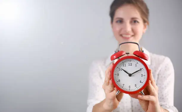 Focus on red alarm clock. Beautiful model in white blouse with curly brunette hair indicating on time. Copy space in left side. Isolated on grey background