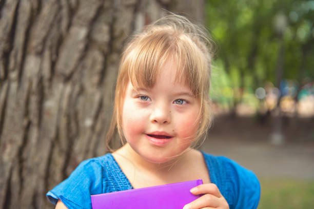 une verticale de trisomie 21 fille d'enfant à l'extérieur ayant l'amusement sur un livre de lecture de parc - child cheerful little girls down syndrome photos et images de collection