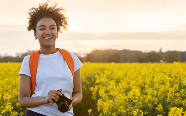 Photo of Beautiful happy mixed race African American girl teenager female young woman smiling outdoors with perfect teeth taking photographs with a camera in a field of yellow flowers