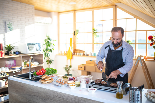 Mature man with apron baking burgers on electrical grill at modern kitchen