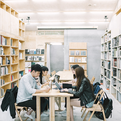 Young students studying together in campus. Classmates are assisting each other in library. Men and women are sitting at desk.