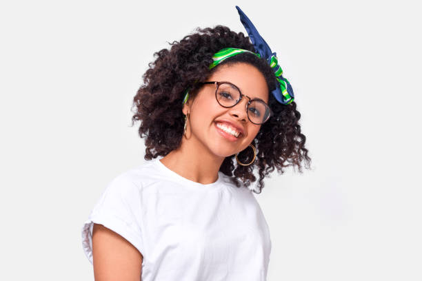 Beautiful happy dark-skinned young woman dressed in white t-shirt, enjoying the weather. African American female smiling broadly, wearing round eyewear posing over white studio wall stock photo