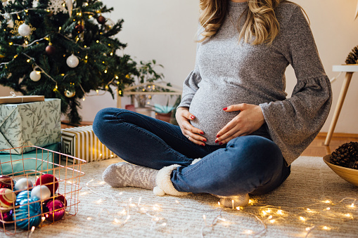 Pregnant woman sitting on floor next to the Christmas tree and holding hands on her stomach