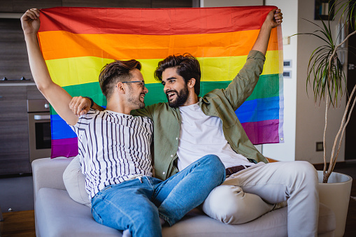 a gay couple is sitting on the couch in the house holding a gay rainbow flag