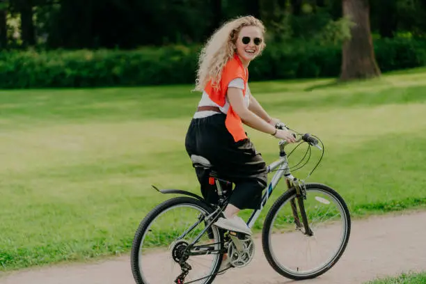 Outdoor image of happy woman rides bike on path, green park around, wears sunglasses, t shirt, pants and sportshoes, has summer holiday, spends spare time actively. People, healthy lifestyle