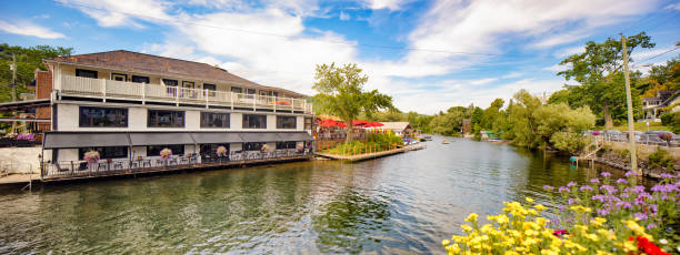 Panoramic view of North Hatley's Massawippi river shore with a restaurant to the left, some people using a pedal boat to the middle and some flowers, parked cars and electric pole to the right.
