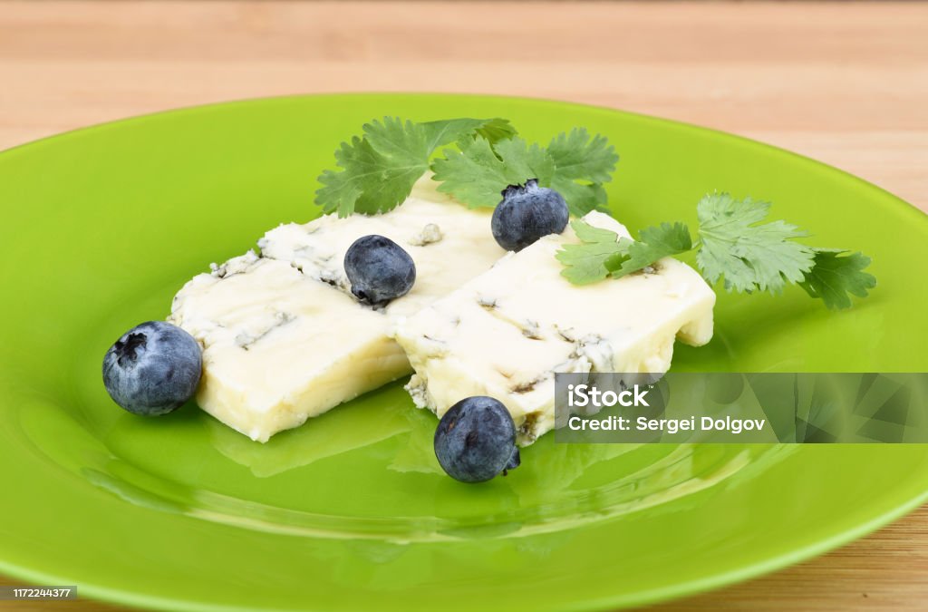 Blueberries and cilantro leaves on slices of delicious blue mold cheese on a green plate. Blueberries and cilantro leaves on slices of delicious blue mold cheese on a green plate. Plate with cheese on a wooden table. Rustic style. Blueberry Stock Photo