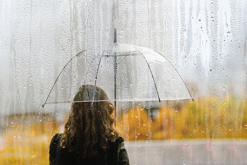 A woman silhouette with transparent umbrella through wet window with drops of rain. Autumn