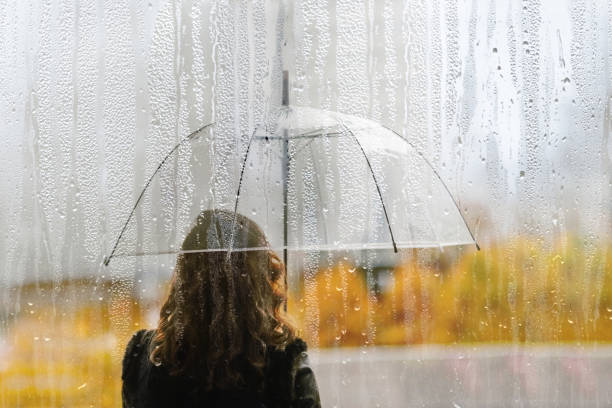 A woman silhouette with transparent umbrella through wet window with drops of rain. Autumn stock photo