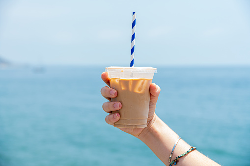 A female's right hand holds a clear plastic cup filled with iced coffee up to the ocean horizon. The top of the cup is parallel with edge of the water. A blue and white striped straw comes out from the top of the plastic cup.
