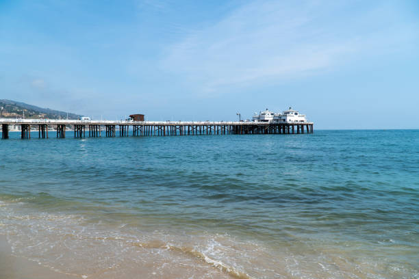 A view of the Malibu pier from the perspective over someone who is stand right where the water washing up onto the sand. It is a clear day with very few clouds in the sky.