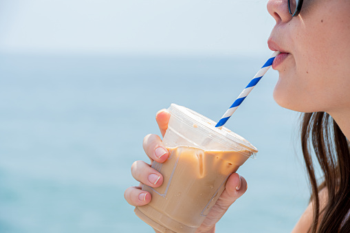 A young college female sips iced coffee from a blue and white striped straw with the ocean horizon in the background. She appears profile to the camera with her lips puckered around the straw and the iced coffee in her right hand.