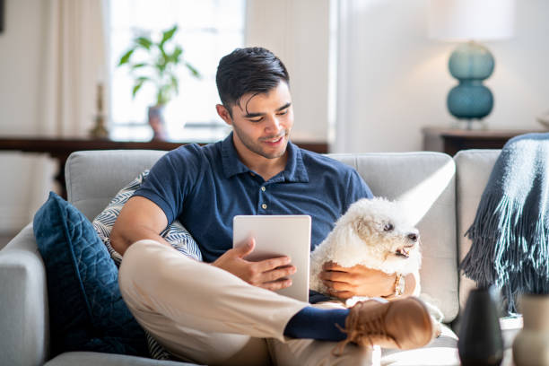młody latynoski mężczyzna siedzący na kanapie ze swoim psem i tabletem - digital tablet men male beauty professional occupation zdjęcia i obrazy z banku zdjęć