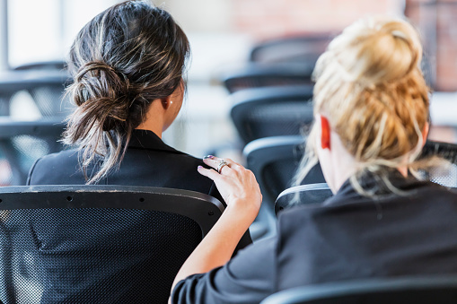 The back of the heads of two multi-ethnic businesswomen sitting in chairs, attending a business conference or seminar.  The focus is on the one with dark hair, a Pacific Islander. The other woman is tapping her on the shoulder trying to get her attention.