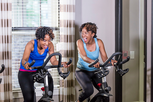 Two mature African-American women in their 40s, identical twins, at the gym exercising on exercise bikes, side by side. They are laughing, encouragements each other, or perhaps having a friendly competition.