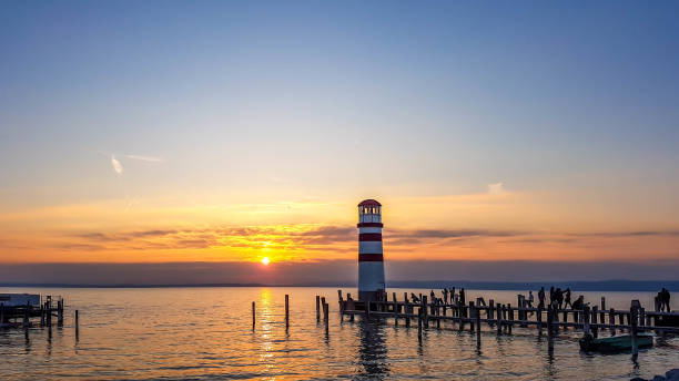 White and red striped lighthouse in Neusiedlersee, on Austrian-Hungarian border. Sun sets over the horizon in the clouds. Sky is orange, lake surface is soft and tender. Few clouds on the sky.