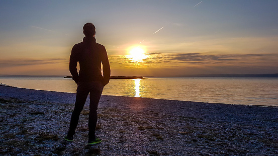 A young man standing on the beach and watching sunset. The beach is pebbly and overgrown with grass. Sun reflects itself in the calm water of the lake. Sunrise above the lake. Freedom and happiness