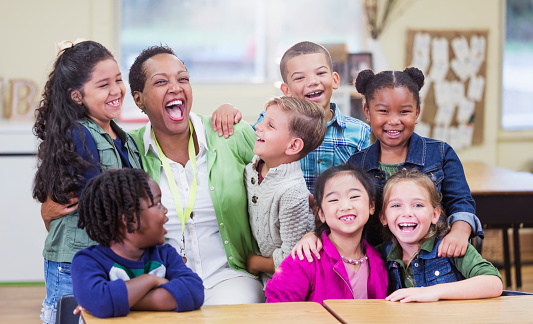 An African-American teacher in an elementary school classroom with seven multi-ethnic boys and girls, 6 and 7 years old. They are having fun, excited to be in school, laughing and smiling. The four school children on the right are looking at the camera. They are in first grade or second grade.