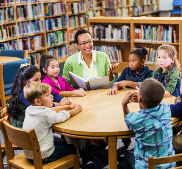 Teacher reading to children in library A multi-ethnic group of seven children, 6 and 7 years old, sitting at a round table in the library with their teacher, a mature African-American woman, who is reading a book to them. librarian stock pictures, royalty-free photos & images