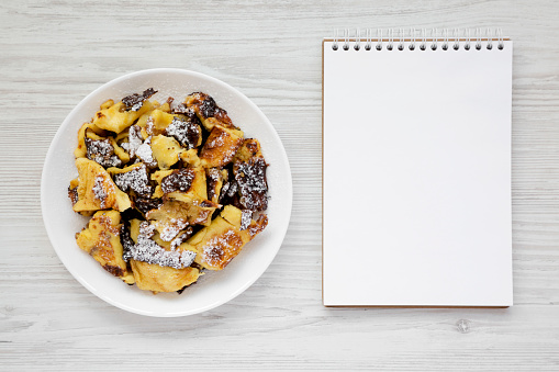 Homemade german Kaiserschmarrn pancake on a white plate, blank notepad on a white wooden surface, top view. Flat lay, overhead, from above. Copy space.