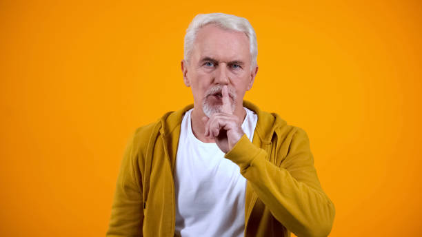 istock Grey-haired pensioner showing silence gesture against orange background, secret 1172311083
