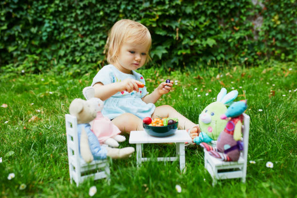 очаровательная маленькая девочка, играющая с мягкими игрушками в парке или саду и делающая их ужин с игрушками и овощами - baby carrot snack healthy eating small стоковые фото и изображения