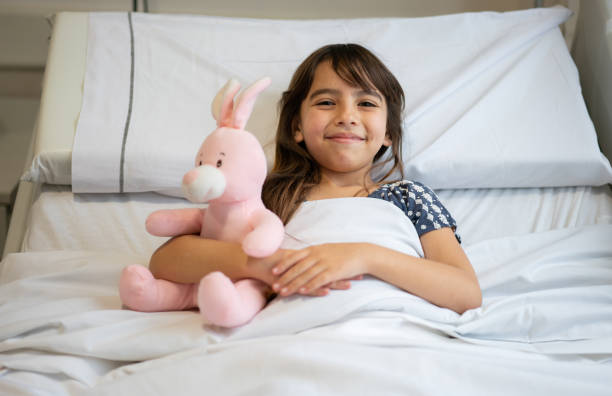 Portrait of beautiful little girl hugging her pink rabbit while lying down on hospital bed smiling at camera Portrait of beautiful little girl hugging her pink rabbit while lying down on hospital bed smiling at camera - Healthcare concepts sick bunny stock pictures, royalty-free photos & images