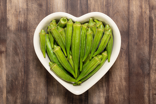 Fresh organic green okra in a heart shaped bowl isolated on a red wood board.