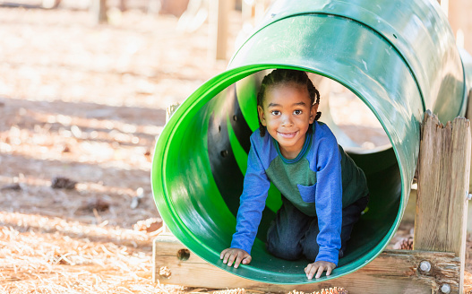 A 4 year old mixed race Hispanic and African-American boy with cornrow braids having fun playing on a playground, crawling through a large tube. He is inside the play equipment, smiling and looking at the camera.