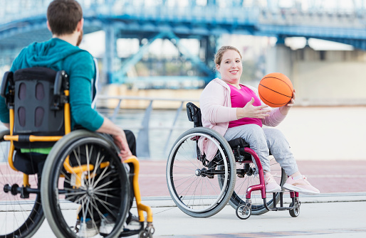 A couple in wheelchairs having fun playing basketball. The young woman, in her 20s, has spina bifida and her mate, a mid adult man in his 30s, has cerebral palsy.  The focus is on the woman who is holding the ball.