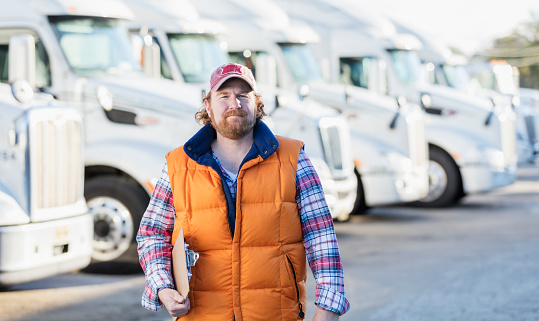 A mature man in h is 40s standing in front of a fleet of semi-trucks or tractor-trailers, holding a clipboard, with a serious, confident expression. He is an experienced truck driver or the owner of manager of a trucking company.