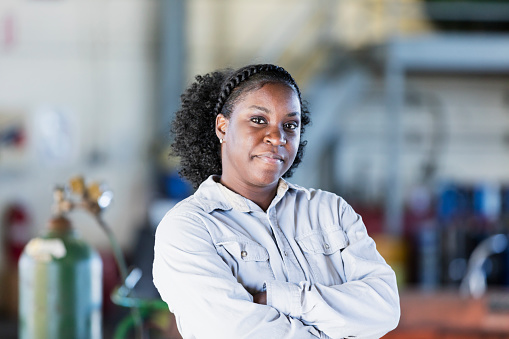 A mid-adult African-American woman in her 30s working in a repair shop. She is standing in the workshop looking at the camera with her arms crossed, a confident expression on her face.