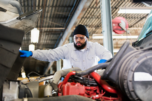 An African-American man in his 30s repairing a semi-truck. The mechanic is looking under the hood of the truck, examining the engine.