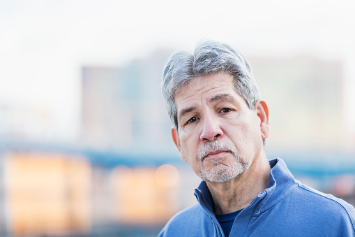 The face of a senior Hispanic man in his 60s, looking at the camera with a serious expression. He looks a bit sad and tired.