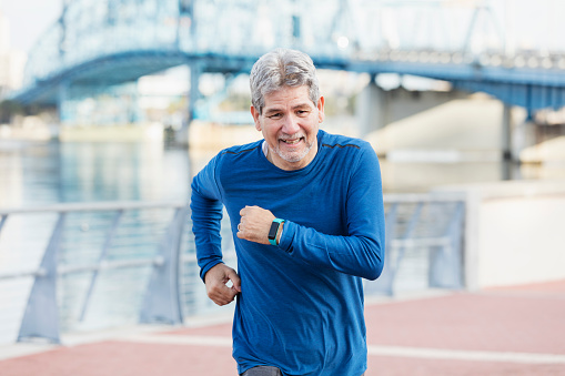 A senior Hispanic man in his 60s exercising with a fitness tracker on his wrist. He is running along a city waterfront. He is smiling, looking at the camera.