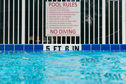 Pool rules sign posted on a fence next to the deep end of a swimming pool. The water depth is posted on tiles at the edge of the pool.