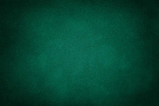 Dark green matte background of suede fabric, closeup. Velvet texture of seamless deep emerald leather. Felt material macro with vignette.