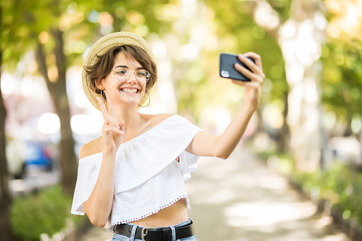 Portrait of young beautiful woman walking in the park take a selfie by camera with peace gesture.